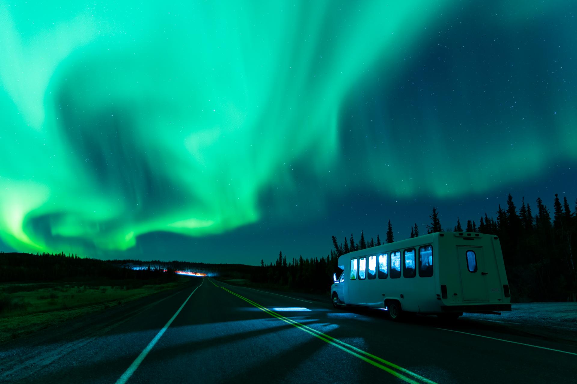 A bus driving on a road at night underneath the Northern Lights.
