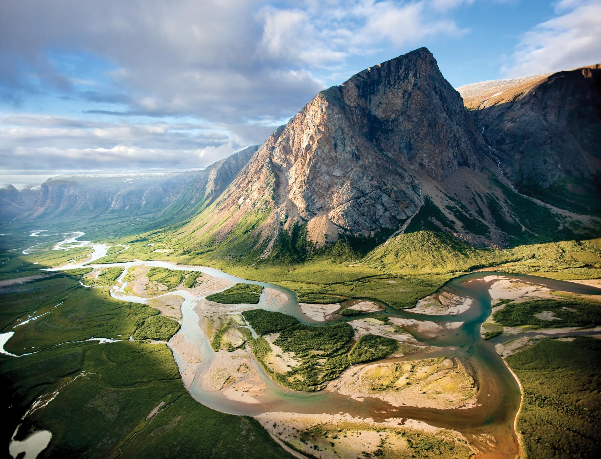 A dramatic aerial view of a steep rocky peak in the Torngat Mountains rising above a braided river winding through green valley wilderness in northern Labrador