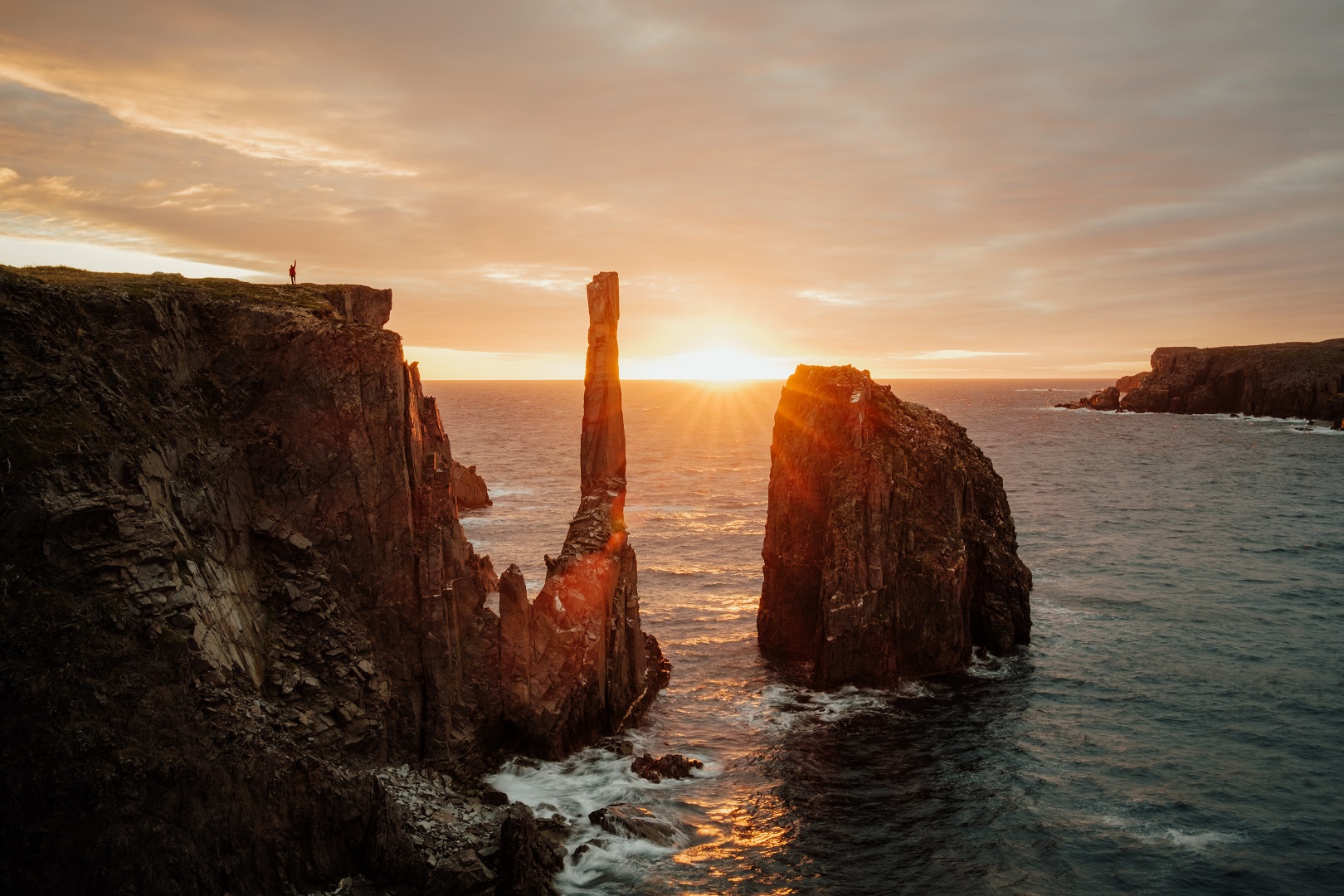 Au coucher du soleil, une personne se tient le bras levé sur une falaise qui donne sur l’océan.