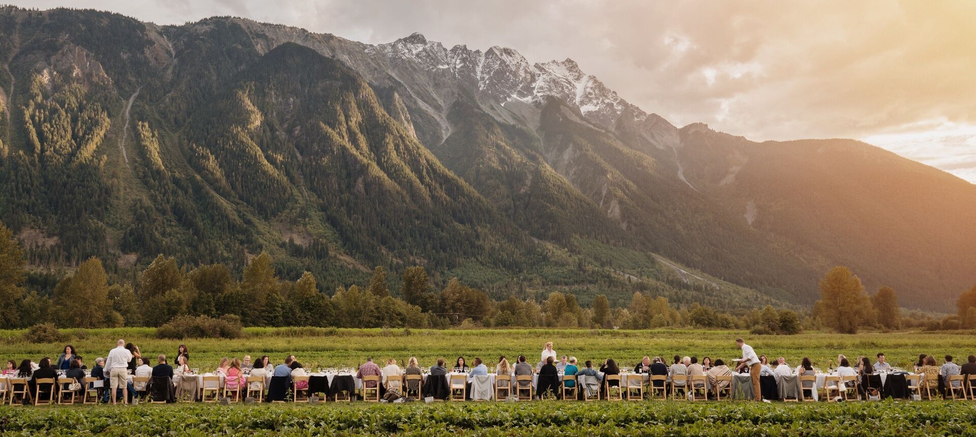 Diners seated at long tables enjoying an outdoor banquet at the base of a mountain