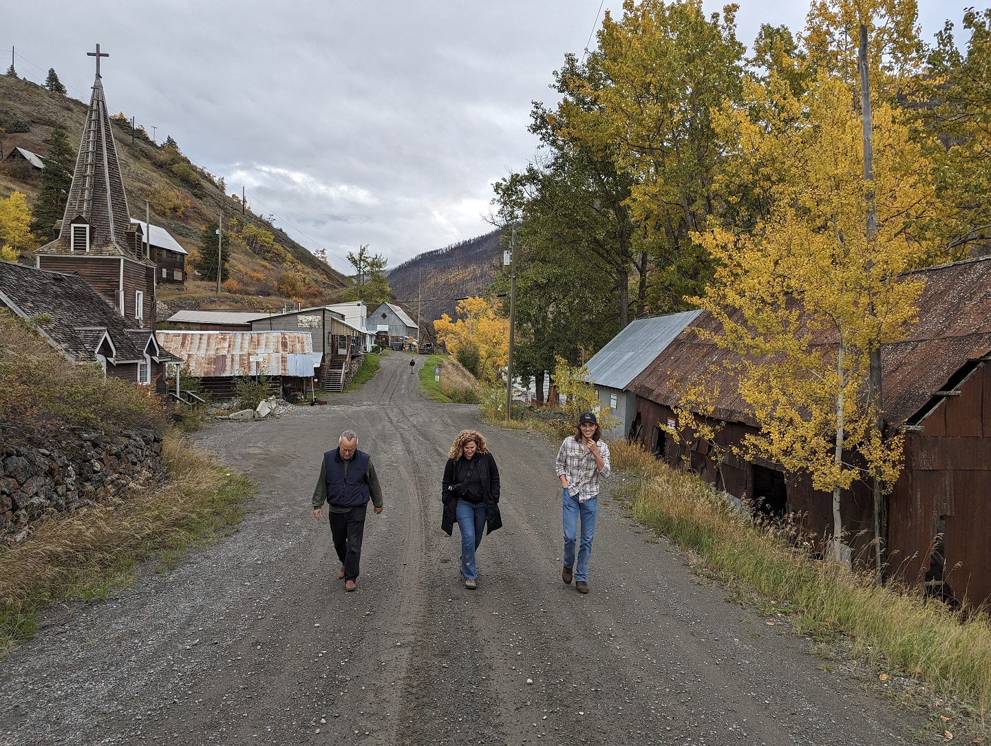 Three people walk on a gravel road flanked by fall foliage.