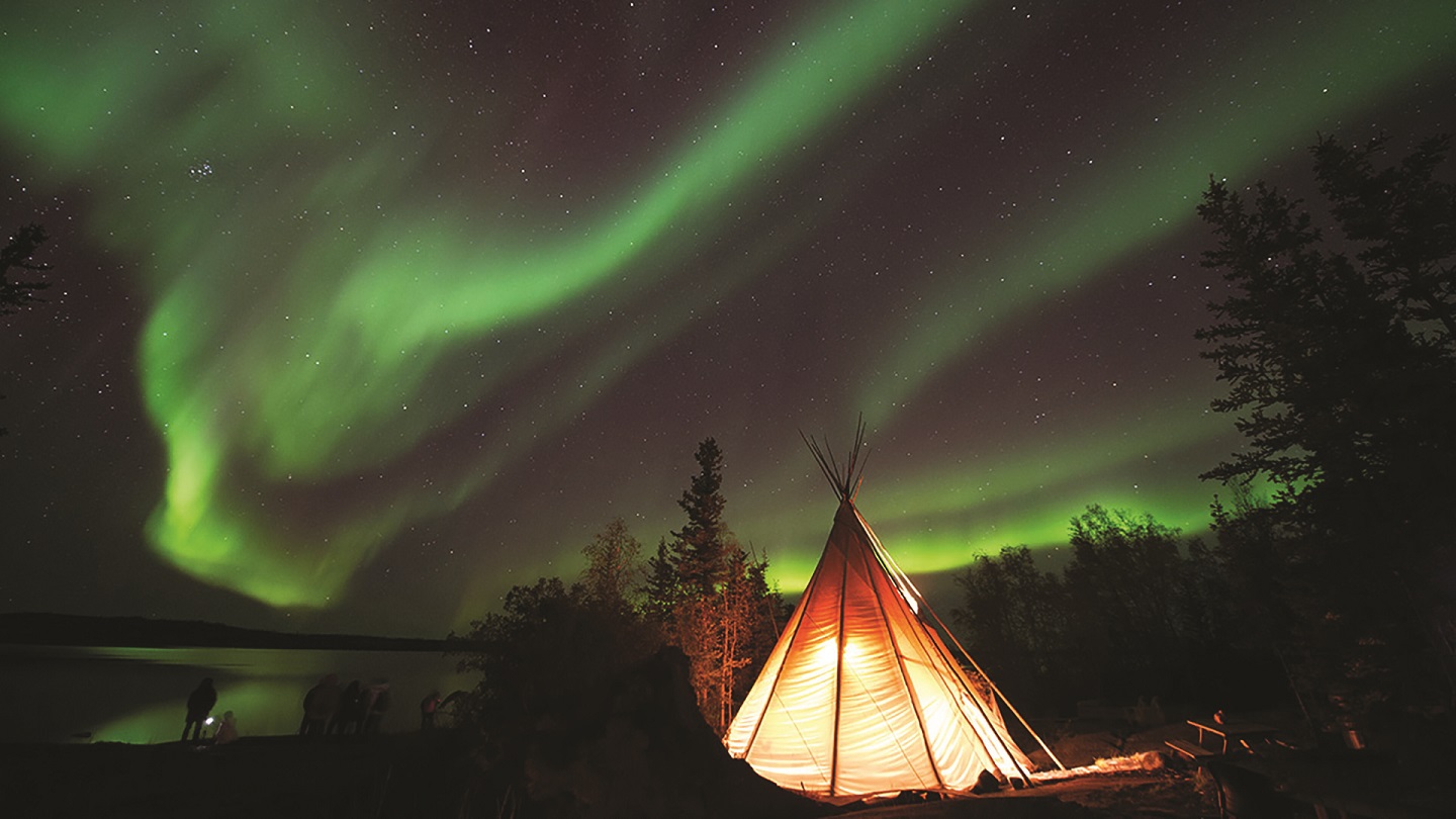 A group of people stand near a lake and next to a teepee, taking photos of the Northern Lights.