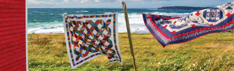 Two colorful quilts are hanging on a clothesline outside, blowing in the wind, with a red building on the left, a grassy field, and a view of the ocean waves and distant coastline in the background.