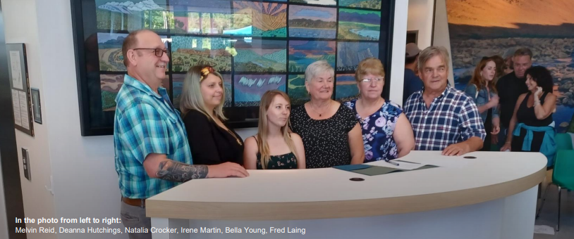 Six people are standing together in front of a reception desk, smiling at the camera, with a colorful artwork displayed on the wall behind them. They are identified as Melvin Reid, Deanna Hutchings, Natalia Crocker, Irene Martin, Bella Young, and Fred Laing from left to right.