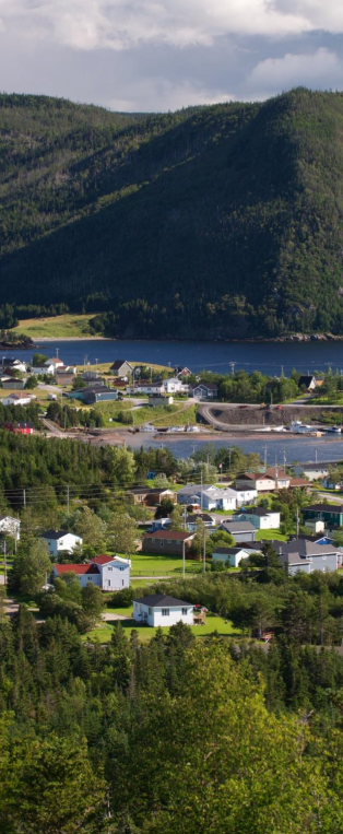 A scenic view of a small town with houses nestled among trees, situated by a river with a bridge, and surrounded by green hills and mountains.