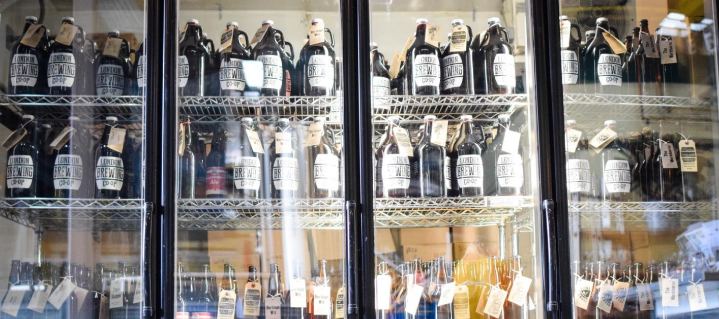 Shelves stocked with glass growlers and bottles of beer from the London Brewing Co-op, displayed inside a refrigerated case.