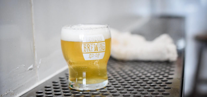 A glass of beer with a foamy head is placed on a perforated metal surface, with "London Brewing Co-op" written on the glass, and a blurred background.