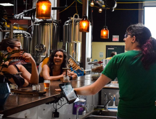 Three people are at a bar inside a brewery, two sitting and chatting with drinks, and one behind the counter with brewing tanks in the background.