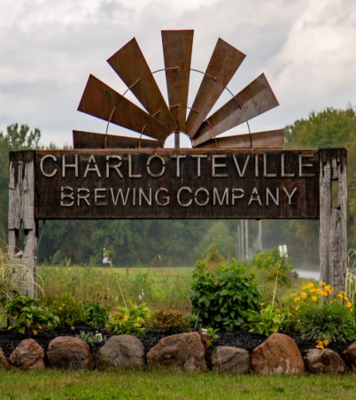 A rustic sign for Charlottesville Brewing Company, featuring metal windmill blades on top, set in a garden with rocks and plants, against a backdrop of trees and a cloudy sky.