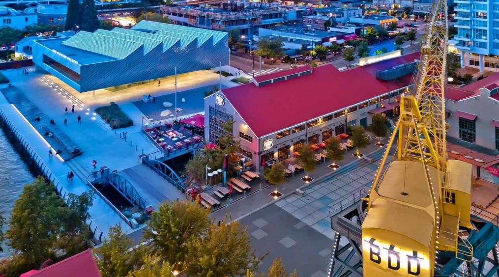 Aerial view of a vibrant waterfront with a modern building, red-roofed restaurants, a yellow crane, and a pedestrian promenade, set in an urban environment.