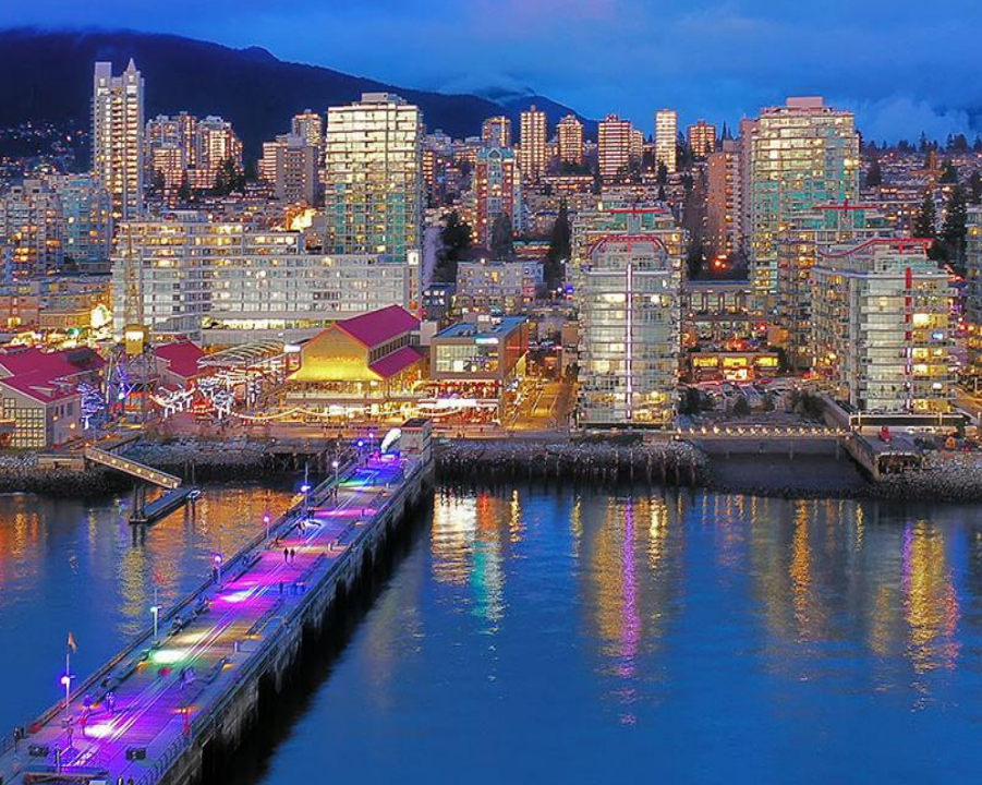 A vibrant waterfront cityscape at night with illuminated docks and a backdrop of tall buildings and mountains.