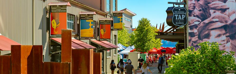 An alley with modern industrial buildings, colorful signs, and a sign for "The Original Tap Barrel," with people walking towards a market area.