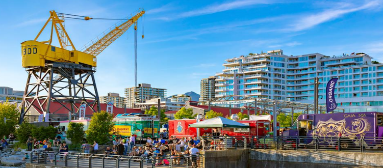 People enjoying a sunny day at a waterfront area with food trucks, a large yellow crane, and modern buildings in the background.