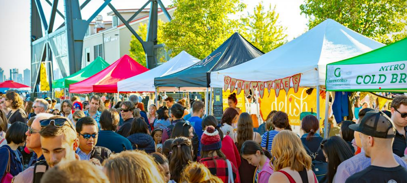 A bustling outdoor market with colorful tents, food stalls, and a diverse crowd, set against an urban backdrop with trees and metal structures.