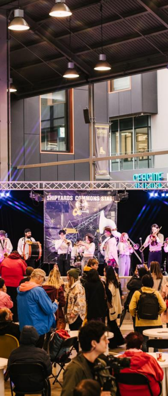 A lively indoor event with a crowd watching a band perform on the "Shipyards Commons Stage."