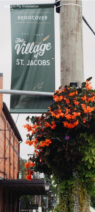 The image shows a street pole banner reading "Rediscover The Village of St. Jacobs, Est. 1852" with a decorative hanging basket of vibrant orange flowers below it.