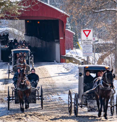 A line of horse-drawn buggies crosses a snowy road and enters a covered bridge in a rural winter scene.