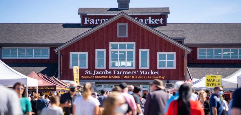 A bustling crowd gathers in front of the red St. Jacobs Farmers' Market building on a sunny day.