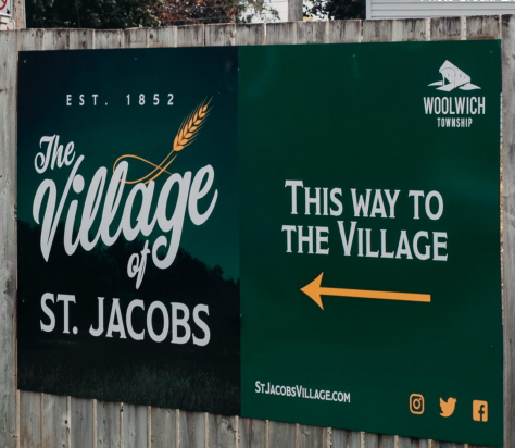 The image shows a sign for "The Village of St. Jacobs," established in 1852, with an arrow pointing the way to the village, and includes logos for Woolwich Township and social media icons.