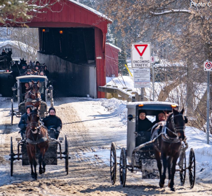 The image shows horse-drawn buggies carrying passengers through a red covered bridge on a snowy road, with a sign indicating to yield to oncoming traffic.