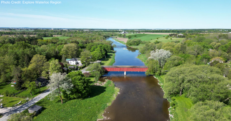 The image shows an aerial view of a picturesque rural landscape with a red covered bridge spanning a calm river, surrounded by lush green trees and fields.