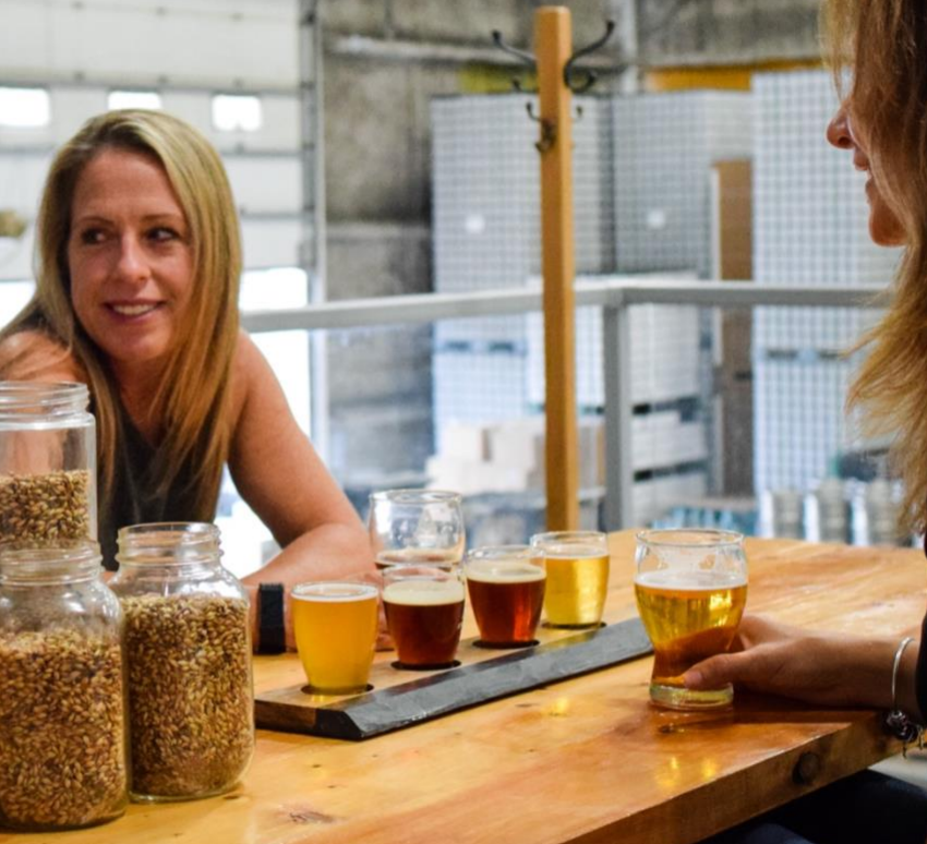 Two people are sitting at a wooden table in a brewery, with jars of grains and a flight of various beers in front of them.