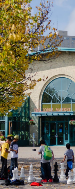 A photo shows children playing a giant chess game in front of the entrance to The Forks Market in Winnipeg, with trees displaying autumn foliage.