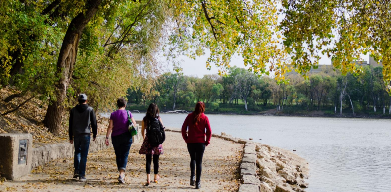 A photo shows four people walking along a scenic riverside path lined with trees and autumn leaves at The Forks in Winnipeg.