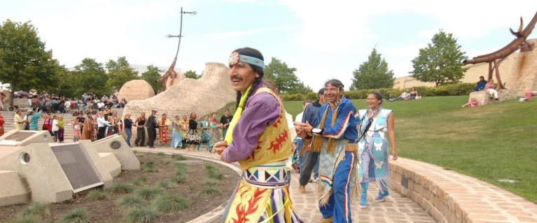 A photo shows Indigenous people in traditional attire dancing and celebrating in an outdoor cultural space at The Forks in Winnipeg, with onlookers and various sculptures in the background.