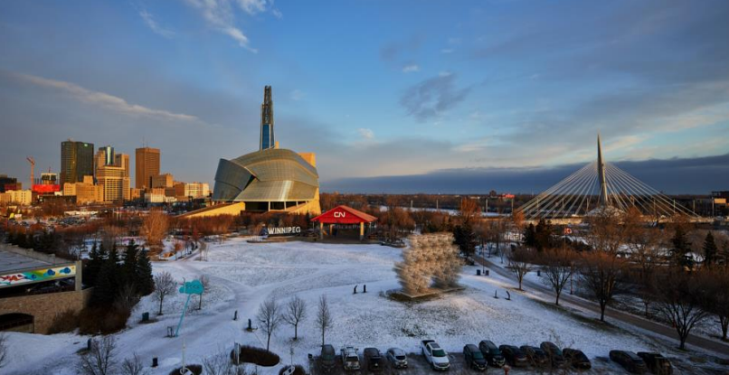 A modern photo shows The Forks area in Winnipeg with the Canadian Museum for Human Rights, the CN Stage, the Esplanade Riel pedestrian bridge, and a skyline of downtown buildings, all lightly dusted with snow.