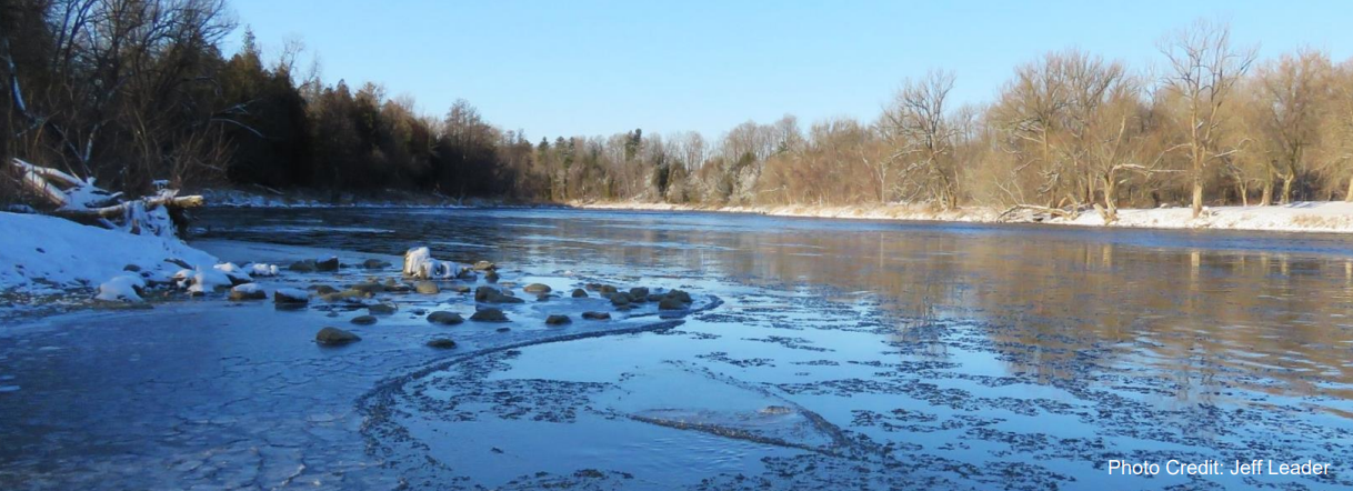 A photo shows a partially frozen river with ice patches, surrounded by snow-covered banks and leafless trees under a clear blue sky, credited to Jeff Leader.