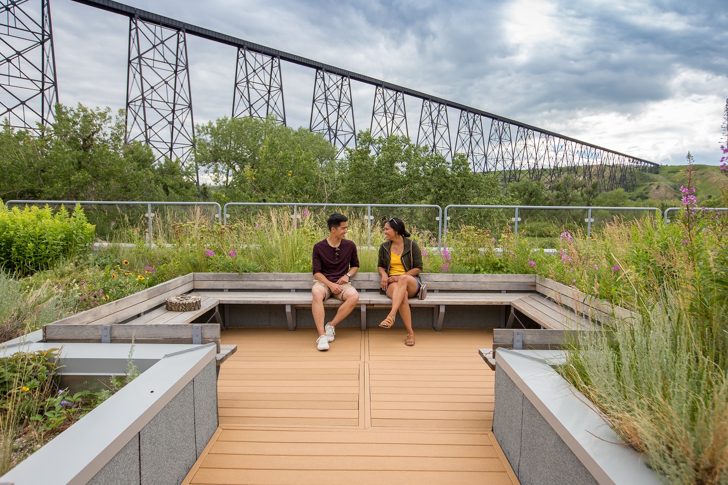 Two people sit on a bench surrounded by plants, with a railway bridge in the background.