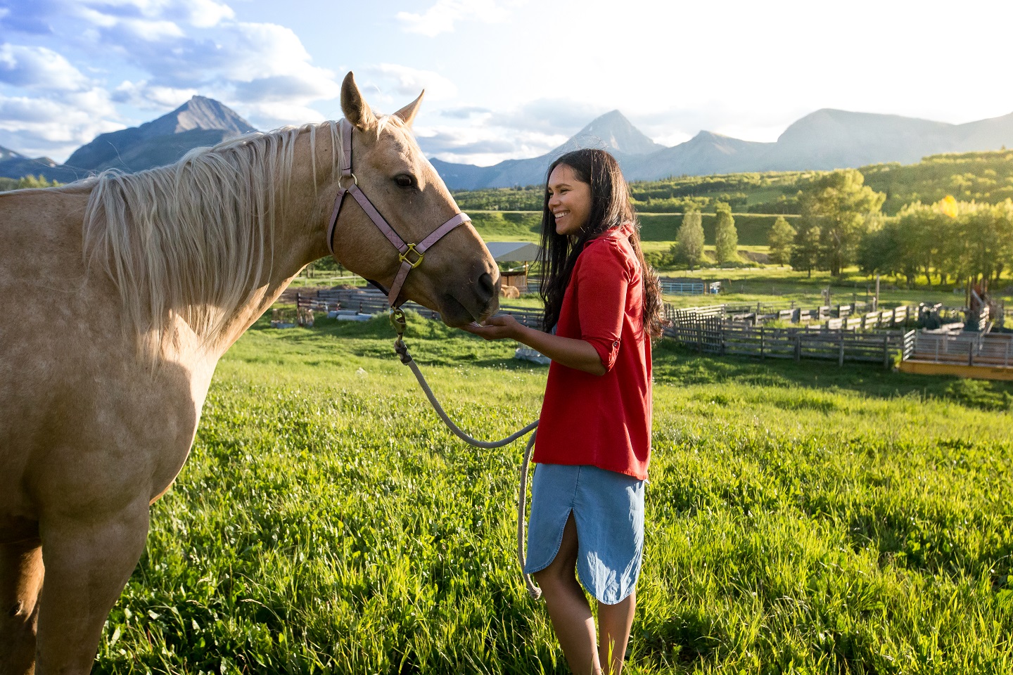 A person smiling, petting a horse in a ranch with mountains in the background.