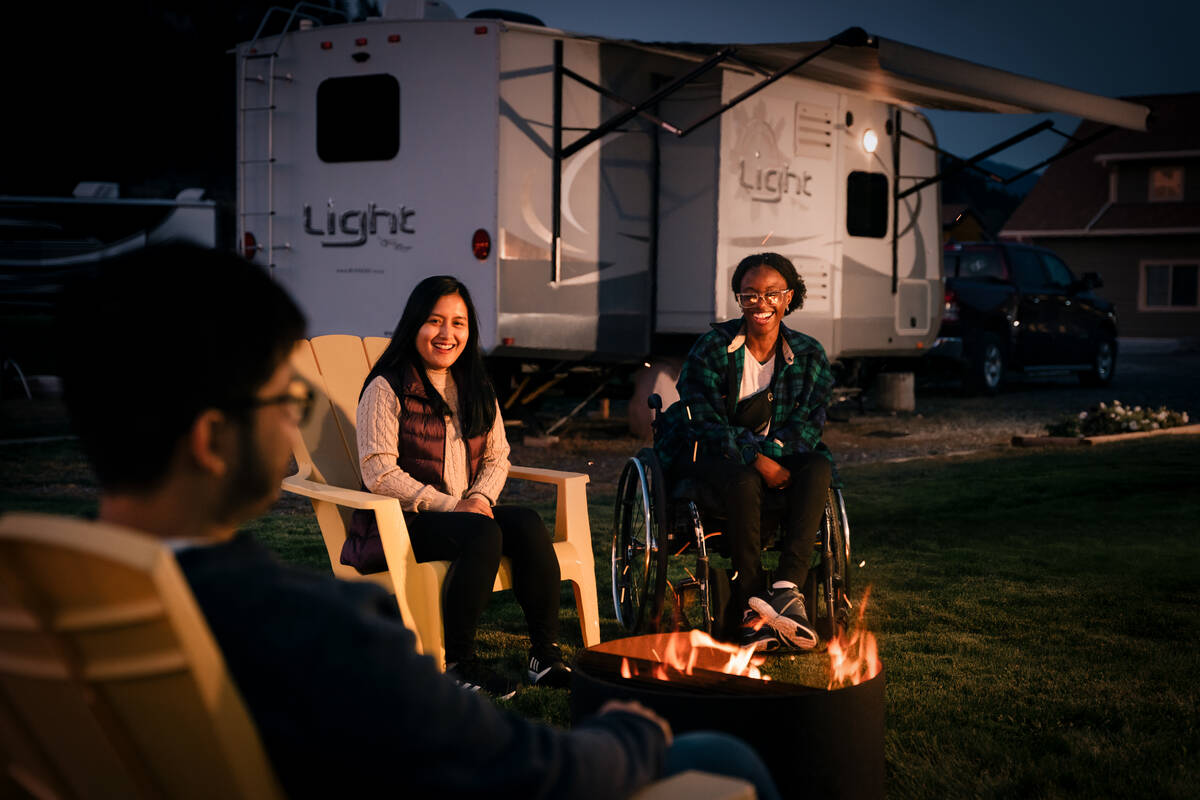 Three people sit by a campfire in the evening, with an RV in the background. 