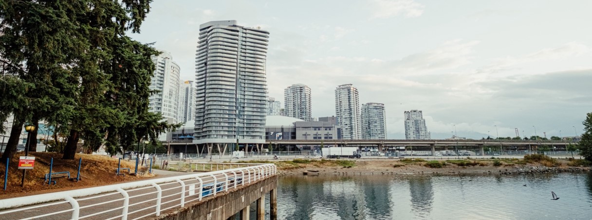 A waterfront view of modern high-rise buildings in Vancouver, BC, with a tree-lined pathway and white railing in the foreground.