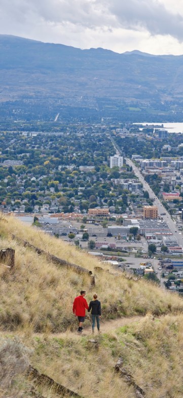 Two people walk along a grassy hillside trail overlooking a sprawling cityscape and distant mountains.