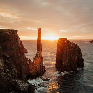 A person raising their arm, standing on a cliff overlooking the ocean at sunset.